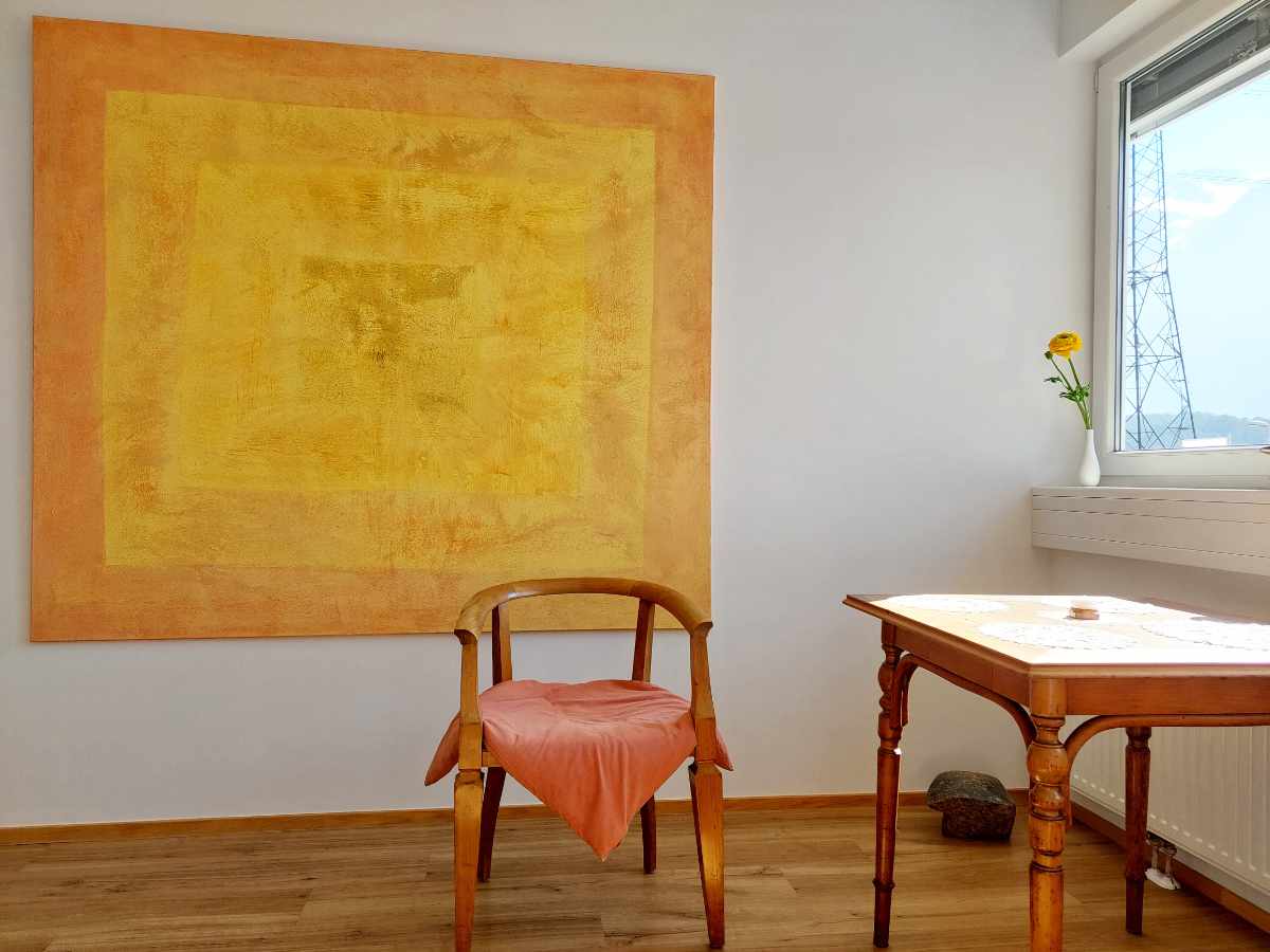 Bild der Praxis von Veronika Falbesoner, Psychologin. Ein Stuhl auf Parkettboden, dahinter ein schönes, großes, orange-gelbes Bild an der Wand. Sonnenlicht scheint durch das Fenster.