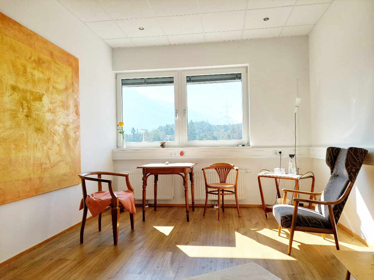 Bild der Praxis von Veronika Falbesoner, Psychologin. Stühle auf Parkettboden, links ein schönes, großes, orange-gelbes Bild an der Wand. Sonnenlicht scheint durch das Fenster.